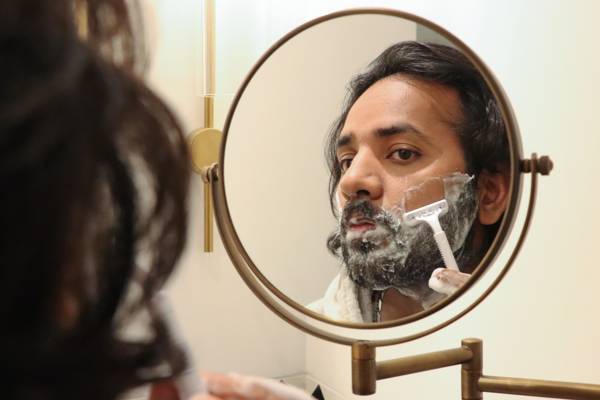 Image of reflection in mirror of Indian man stood at sink bathroom wearing bathrobe using razor to trim facial hair, cutting beard and moustache, self care and grooming concept